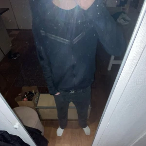 Svart Nike tech Hoodie - Säljer en svart hoodie med dragkedja och huva. Den har en enkel design med långa ärmar och en bekväm passform. Perfekt för en avslappnad stil.