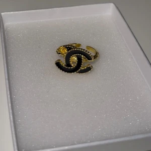 Guldfärgad ring med svart detalj - Elegant guldfärgad ring med en svart detalj i form av ett stiliserat mönster. Perfekt för att ge en lyxig touch till din outfit. Ringen har en justerbar design för att passa olika storlekar. Helt ny och oanvänd. 
