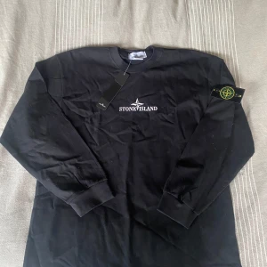 Svart tröja från Stone Island - Säljer en svart långärmad tröja från Stone Island i 100% kammad bomull. Tröjan har en stor logga på ryggen och en ikonisk patch på ärmen. Perfekt för en stilren och avslappnad look.