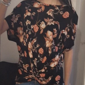 Blommig topp med knutdetalj - Säljer en svart topp med vackert blommönster i rosa och beige. Toppen har korta ärmar och en snygg knutdetalj framtill. Är i storlek S 💕