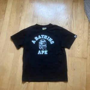 Svart t-shirt från A Bathing Ape - Snygg svart t-shirt från A Bathing Ape med det ikoniska logotyptrycket på framsidan. T-shirten har korta ärmar och är tillverkad i mjuk bomull. Perfekt för en avslappnad stil.