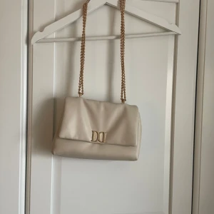 Beige axelväska  - Elegant beige axelväska med en stilren guldkedja och dekorativt emblem på framsidan. Väskan är köpt från glitter för 400 kr. Väskan passar superbra till allt och är jättesnygg 🤗🤗☺️