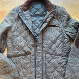 Grön quiltad jacka från Vedoneire - Säljer en snygg grön quiltad jacka från Vedoneire. Jackan har en stilren design med dragkedja och knappar framtill. Den har långa ärmar och flera praktiska fickor med bruna detaljer. Perfekt för höst och vår! Nypris 1500kr