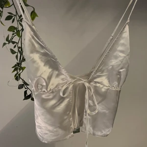 Vit satinlinne med knytband - Elegant vit satinlinne med smala axelband och knytband framtill, korsade band i ryggen. perfekt till varma sommardagar, festivaler och fester! ser väldigt stilren ut och kan göra en basic outfit till något snyggt. aldrig använd och prislapp kvar! den är storlek M men skulle säga att den mer är som en xs/s! skriv ifall du har fler frågor💗