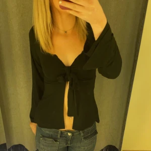 Svart långärmad topp från Design by Si - Snygg svart långärmad topp från Design by Si med knytning framtill. Perfekt för en stilren look. Toppen har en v-ringad design som ger en elegant touch.