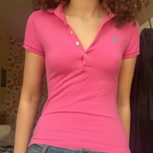 Rosa pikétröja från Ralph Lauren - Säljer en snygg rosa pikétröja från Ralph Lauren med klassisk krage och knappar framtill. Tröjan har en liten blå logga på bröstet och korta ärmar. Perfekt för en avslappnad stil.
