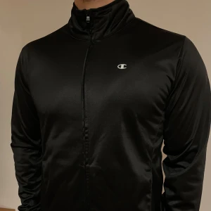 Svart ziptröja från Champion - Säljer en svart träningsjacka från Champion med dragkedja framtill. Jackan har en stilren design med långärmade ärmar och en liten logotyp på bröstet. Perfekt för träning eller som en lätt överdragströja. Passar en S/M
