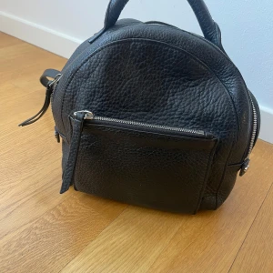 Svart ryggsäck i skinn - Snygg svart ryggsäck i skinn med dragkedja framtill och justerbara axelremmar. Perfekt för en stilren look och praktisk för vardagsbruk. Den har en framficka med dragkedja för extra förvaring.