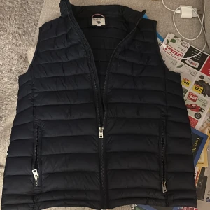 Svart dunväst från U.S. Polo Assn. - Snygg svart dunväst från U.S. Polo Assn. med dragkedja framtill och två praktiska fickor med dragkedjor. Perfekt för kyligare dagar och ger en stilren look.