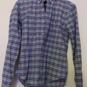 Rutig skjorta från Polo Ralph Lauren - Säljer en snygg rutig skjorta från Ralph Lauren i blått, rött och vitt. Skjortan har en broderad logga på bröstet och knappar framtill. Den är slim fit och långärmad, perfekt för en stilren look.