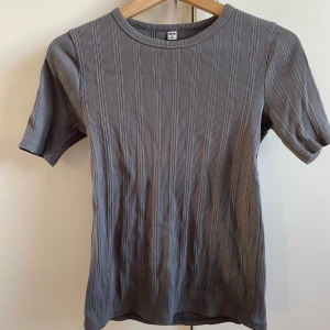 Grå ribbad t-shirt från Uniqlo - Säljer en stilren grå ribbad t-shirt från Uniqlo. T-shirten har en normal passform med lite längre kortärmar och rund halsringning. Perfekt för en avslappnad look men med en elegant touch. 