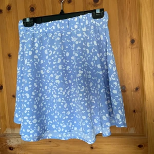 Ljusblå kjol med mönster från Monki - Säljer en söt ljusblå kjol från Monki med ett vitt abstrakt mönster. Kjolen har en bekväm resår i midjan och är perfekt för en avslappnad stil. Den är lätt och luftig, vilket gör den idealisk för varmare dagar.