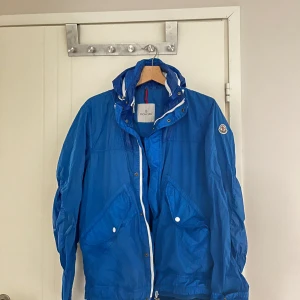 Blå vindjacka från Moncler - Säljer en snygg blå vindjacka från Moncler med dragkedja och knappar. Jackan har en broderad Moncler-logga på ärmen och en justerbar huva. Perfekt för blåsiga dagar!