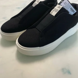 Svarta sneakers från Lager 157 - Snygga svarta sneakers från Lager 157 med en stilren design. Skorna har en vit sula och är perfekta för en casual look. De är märkta med storlek 38 och har en enkel slip-on stil utan snörning.