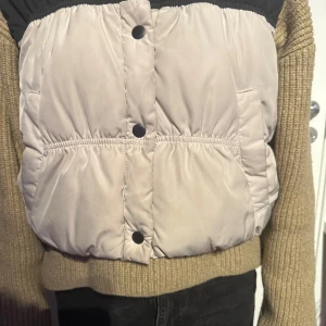 Beige och svart pufferjacka - Säljer en snygg beige och svart pufferjacka med knappar. Jackan har en cool kontrastdesign med svarta detaljer upptill och beige nedtill. Perfekt för att hålla värmen med stil.