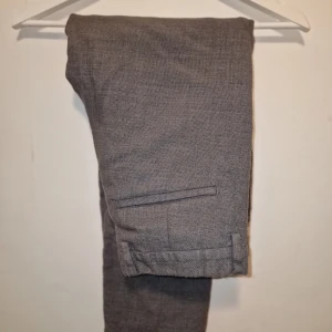 Grå kostymbyxor från COS - Eleganta grå byxor med en klassisk design. Byxorna har en rak passform och är försedda med fickor bak. Perfekt för en stilren och sofistikerad look. Över 1000 kr vid inköp.  