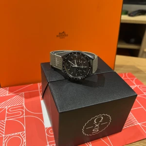 Svart klocka från Omega x Swatch - Snygg svart klocka från Omega x Swatch med grått armband. Klockan har en tydlig urtavla med vita detaljer och en tachymeter-skala runt ringen. Perfekt för den stilmedvetna som vill ha något unikt på handleden.(mission to mercery)kvitto och box finns.