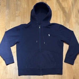 Navy blue hoodie från Ralph Lauren - Säljer en stilren navy blue hoodie från Ralph Lauren med dragkedja och klassisk logga på bröstet. Perfekt för en avslappnad look. Hoodien har långa ärmar och en praktisk huva med snörning.