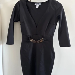 Svart klänning från NLY One - Snygg svart klänning från NLY One med v-ringning och trekvartsärmar. Den har en gulddetalj i midjan som ger en extra touch. Perfekt för en kväll ute eller en speciell tillställning. Storlek XS. Använd 1 gång max så ser ut som ny 