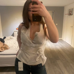 Vit omlott topp från Zara - Snygg vit omlott topp från Zara. Den är gjord i ett glansigt material. Toppen är ärmlös och har en knytning i ryggen. Går att knyta på 2 sätt - bild 1&2 är knytning framtill, bild 3&4 är knytning bak. Är i strl M men kan även användas för större och mindre.