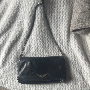 Zadig väska - Snygg svart skinnväska från Zadig & Voltaire med kedjeaxelrem och coolt vingmotiv på framsidan. Väskan har en praktisk dragkedja på baksidan för extra förvaring. Perfekt för att ge din outfit en edgy touch.
