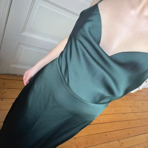 Mörkgrön satin klänning - Elegant mörkgrön klänning med smala axelband och djup urringning. Klänningen har en slät och glansig yta, vilket ger en lyxig känsla. Perfekt för speciella tillfällen. Använt en gång. Jag är 155 cm lång och klänningen når ner till mina fötter.