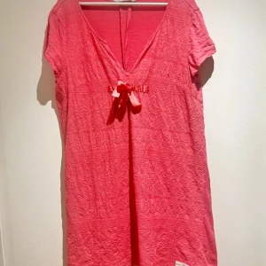 Rosa klänning från Odd Molly - Charmig rosa klänning från Odd Molly med v-ringning och knytband framtill. Klänningen har ett vackert broderat mönster och korta ärmar. Perfekt för en somrig look! Sömmar som gått upp lite vid ena armen