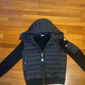 Svart dunjacka från Moncler - Storlek L men sitter som M/L Snygg svart dunjacka från Moncler med huva och dragkedja. Jackan har långa ärmar och ribbade muddar. Perfekt för kyligare väder med sin quiltade design och stilrena look. HELT NY