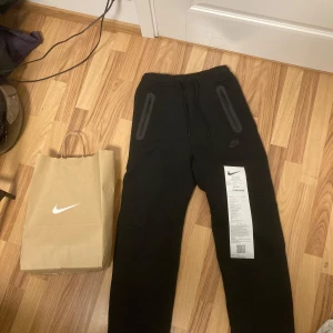 Svarta mjukisbyxor från Nike - Säljer ett par svarta mjukisbyxor från Nike med elastisk midja och två fickor med dragkedja. Perfekta för en avslappnad stil eller träning. Byxorna har en diskret Nike-logga på benet. Kvitto följer med⭐️ 