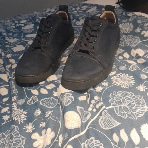 Svarta sneakers från Christian Louboutin - Säljer ett par svarta sneakers från Christian Louboutin med klassisk röd sula. Skorna har rund tå, snörning och är tillverkade i skinn. Perfekta för dig som vill ha något stilrent men ändå sticker ut med den ikoniska röda sulan.