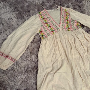 Vit klänning med broderade blommor från Axander - Söt vit klänning med v-ringning och broderade blommor i gult, rött och grönt på bröstet. Långa ärmar med broderade detaljer och lätt rynkad kjol. Perfekt för en somrig look.