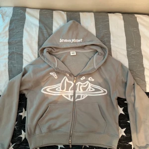 Broken Planet - Säljer en grå hoodie från Broken Planet med vit tryckt design på bröstet och broderad logga på huvan. Tröjan har dragkedja framtill och känguruficka. Perfekt för en chill och avslappnad stil. Helt ny!