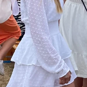 Vit prickig klänning med volang - Superfin vit klänning med prickigt mönster och volangdetaljer. Klänningen har långärmade ärmar och markerad midja, vilket ger en feminin siluett. Perfekt för sommarens alla tillfällen!