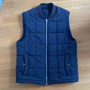 Mörkblå quiltad väst från Riley - Stilren mörkblå quiltad väst från Riley med dragkedja framtill och två fickor med knappstängning. Västen har en klassisk rak passform och ribbad krage. Perfekt för lager-på-lager-stil.