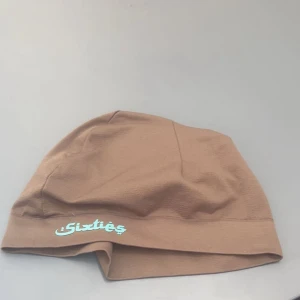 Sixties mössa - Säljer en beige mössa från Sixties med enkel design och tryckt logga framtill. Mössan är mjuk och tunn, perfekt för mildare väder. Passar dig som gillar stilrena accessoarer.