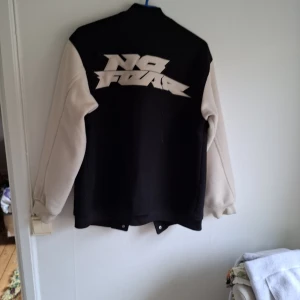 Svart och vit bomberjacka från No Fear - Säljer en svart och vit bomberjacka från No Fear med stora loggor på ryggen och mindre logga på bröstet. Jackan har knappar framtill och två fickor. Ärmarna är vita och kroppen är svart, klassisk college-stil.