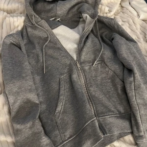 Grå hoodie med dragkedja - Säljer en grå hoodie med dragkedja och huva. Tröjan har fickor framtill och justerbara snören vid huvan. Perfekt för chill dagar eller när du vill ha något bekvämt på dig.