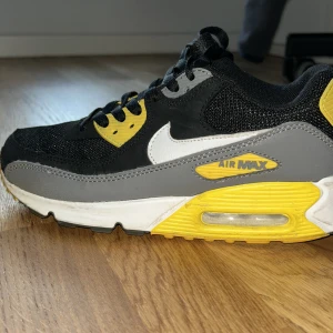 Nike Air Max 90 svart/gul - Nike Air Max 90 sneakers i svart mesh och syntet med vita och grå detaljer samt gula inslag på sula och snörhål. Klassisk vit swoosh på sidan och synlig Air Max-dämpning i sulan. Perfekta för dig som gillar streetstyle.