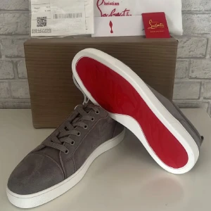 Grå sneakers från Christian Louboutin - Säljer ett par gråa sneakers från Christian Louboutin med klassisk röd sula. Skorna har snörning och rund tå, tillverkade i mocka med vita platta sulor. Perfekta för dig som vill ha en stilren och lyxig look.