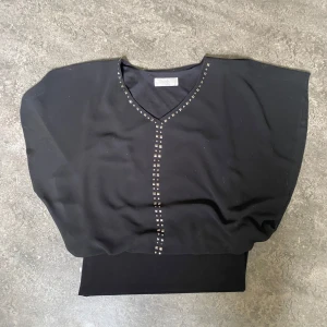 Svart topp med nitar från Wallis - Säljer en svart topp från Wallis Petite i storlek S. Toppen har v-ringning och dekorativa nitar längs halsringningen och mitt fram. Modellen är lös med bred mudd nedtill och korta ärmar. Perfekt för en stilren look.