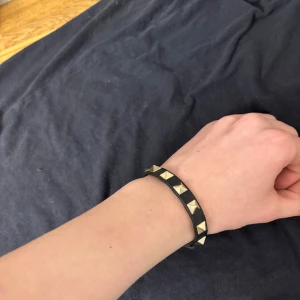 Valentino armband - Säljer ett svart armband från Valentino med guldiga pyramidformade nitar och spänne. Armbandet är gjort i skinn och har justerbar längd med klassiskt Valentino-spänne. Perfekt accessoar för att ge din outfit en edgy touch.