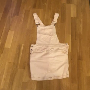 Ljusrosa hängselklänning - Säljer en ljusrosa hängselklänning med justerbara axelband och knappar i sidorna. Klänningen har fickor framtill och är i en kort modell. Perfekt för en avslappnad och trendig look.