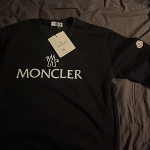 Svart Moncler tröja med logga - Säljer en svart Moncler tröja i storlek M med stor vit logga framtill och klassisk Moncler-patch på ärmen. Tröjan har rund halsringning och är tillverkad i mjukt material. Perfekt för dig som gillar stilrena märkesplagg. Påsen följer med. Pruta gärna 