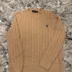 Beige kabelstickad tröja från Polo Ralph Lauren - Kabelstickad beige tröja från Polo Ralph Lauren med rund halsringning och broderad logga på bröstet. Tröjan har långa ärmar och ribbade muddar vid ärmslut och nederkant. Perfekt för en klassisk och stilren look.