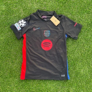 Svart FC Barcelona fotbollströja med Lamine Yamal - Säljer en svart FC Barcelona fotbollströja med röda och blå sidopaneler, klubbmärke på bröstet och stor röd Spotify-logga. Nike-logga på bröstet och Champions League-märke på ärmen. Namn och nummer 'Lamine Yamal 19' på ryggen.