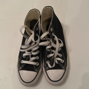 Svarta Converse All Star high-top sneakers - Klassiska svarta Converse All Star high-top sneakers med vit tå och vita snören. Skorna har den ikoniska loggan på sidan och vit sula med svart rand. Perfekta för en avslappnad och cool stil.