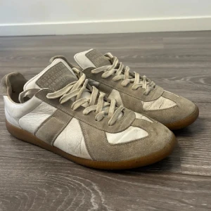 Beige och vita sneakers med mockadetaljer - Säljer ett par klassiska sneakers i beige och vitt med snygga mockadetaljer och snörning. Skorna har en platt gummisula och rund tå, perfekta för en stilren vardagslook.