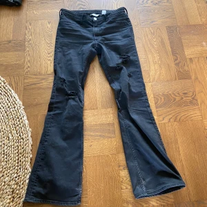 Svarta bootcut jeans med slitningar från H&M - Svarta jeans från H&M med bootcut-ben och slitningar på båda benen. Låg midja (egentligen mid) och klassisk femficksmodell. Jeansen har en cool, avslappnad look och är perfekta för dig som gillar en trendig stil.