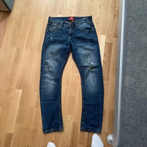 Blå jeans med slitningar - Säljer ett par blå jeans med snygga slitningar och en liten reva på ena benet. Klassisk femficksmodell med raka ben och normal passform. Perfekta för en avslappnad stil.