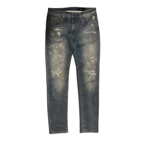 Distressed jeans från Dolce & Gabbana - Säljer ett par blå jeans från Dolce & Gabbana med slitna detaljer och distressed look. Jeansen har klassisk femficksdesign, knappgylf och raka ben. Perfekta för dig som gillar en trendig och avslappnad stil.